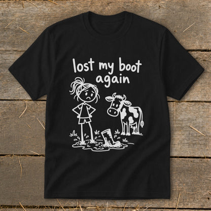 A black t-shirt features white graphics of a sad stick-figure woman standing in mud next to a cow and a boot, with text reading "lost my boot again".