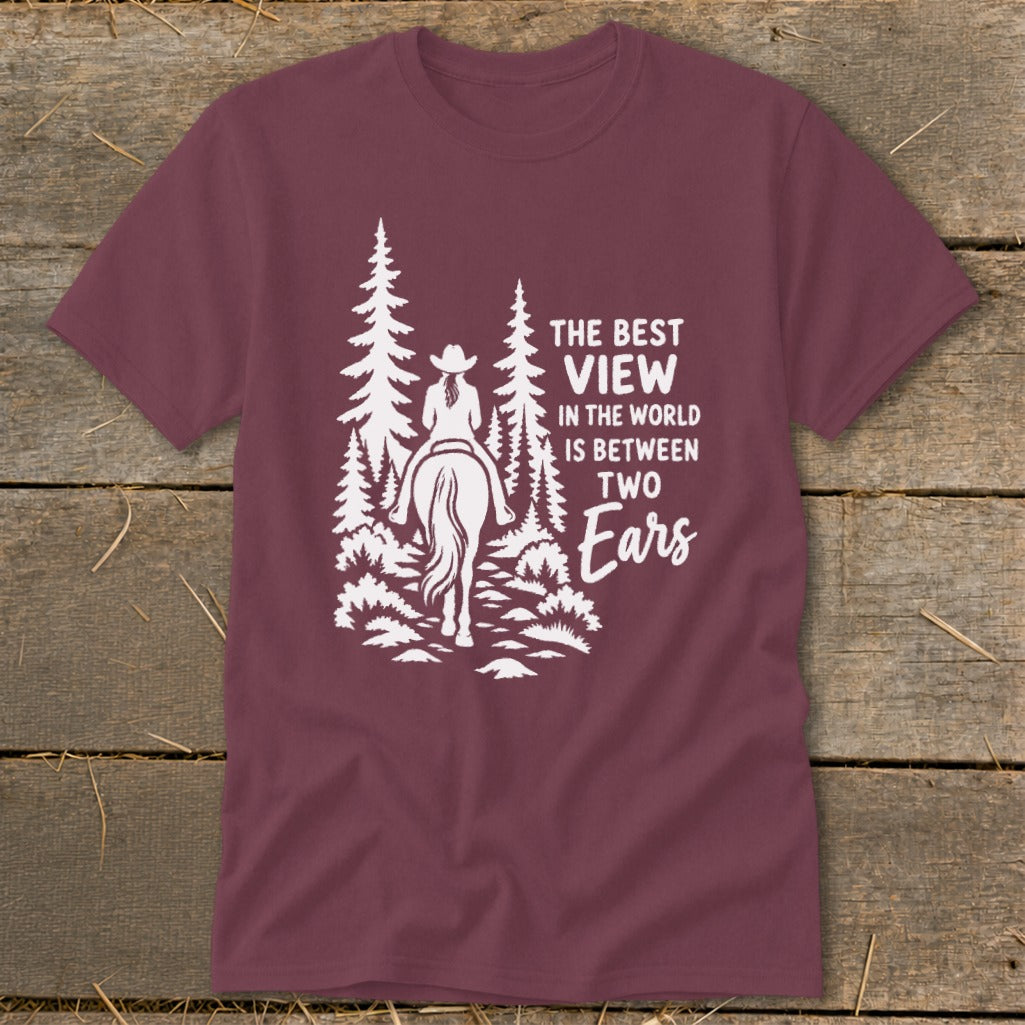 Maroon t-shirt with a white graphic of a horseback rider among pine trees and text reading The best view in the world is between two ears.