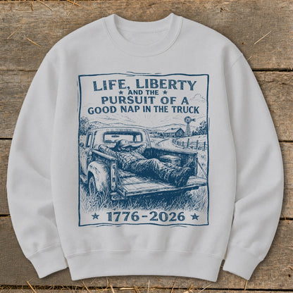 White sweatshirt with graphic of man napping in truck bed, text "Life, Liberty and the Pursuit of a Good Nap in the Truck 1776-2026".