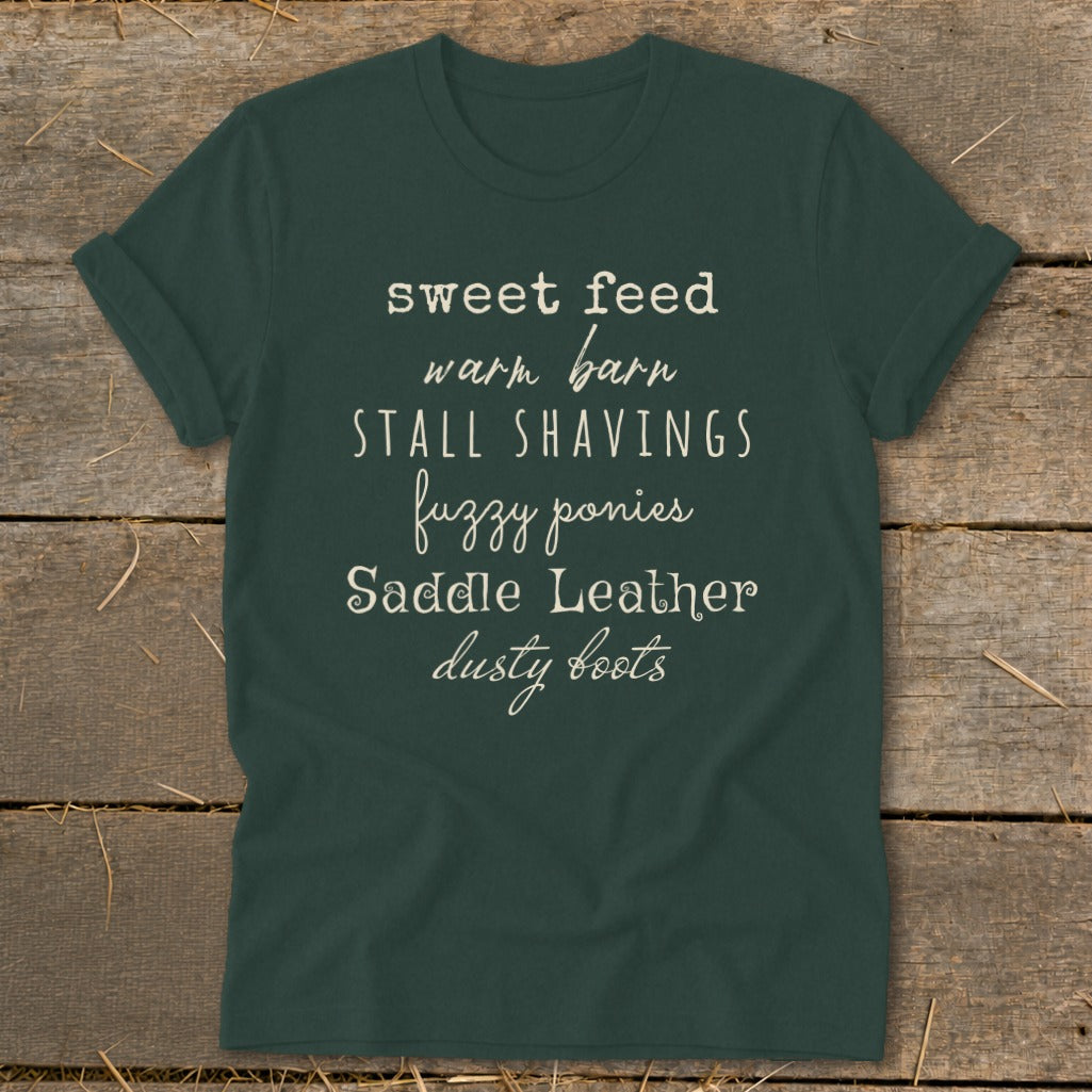 Dark green t-shirt with text: sweet feed, warm barn, stall shavings, fuzzy ponies, saddle leather, dusty boots.