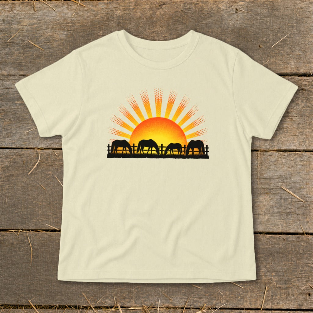 Silhouette of horses grazing in front of a rising sun graphic on a cream t-shirt.