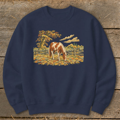 Brown and white horse grazing in a field of autumn leaves with a tree and fence.