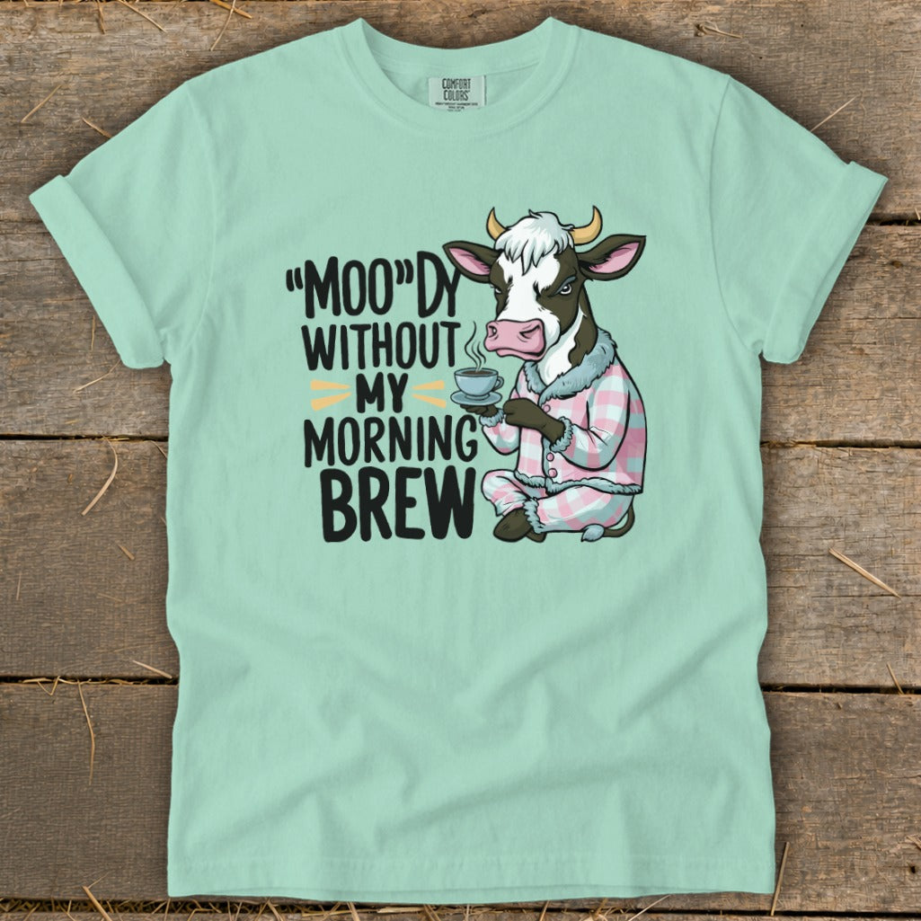 Light mint green t-shirt with a cartoon cow in pajamas holding coffee. Text reads "Moo"dy Without My Morning Brew."