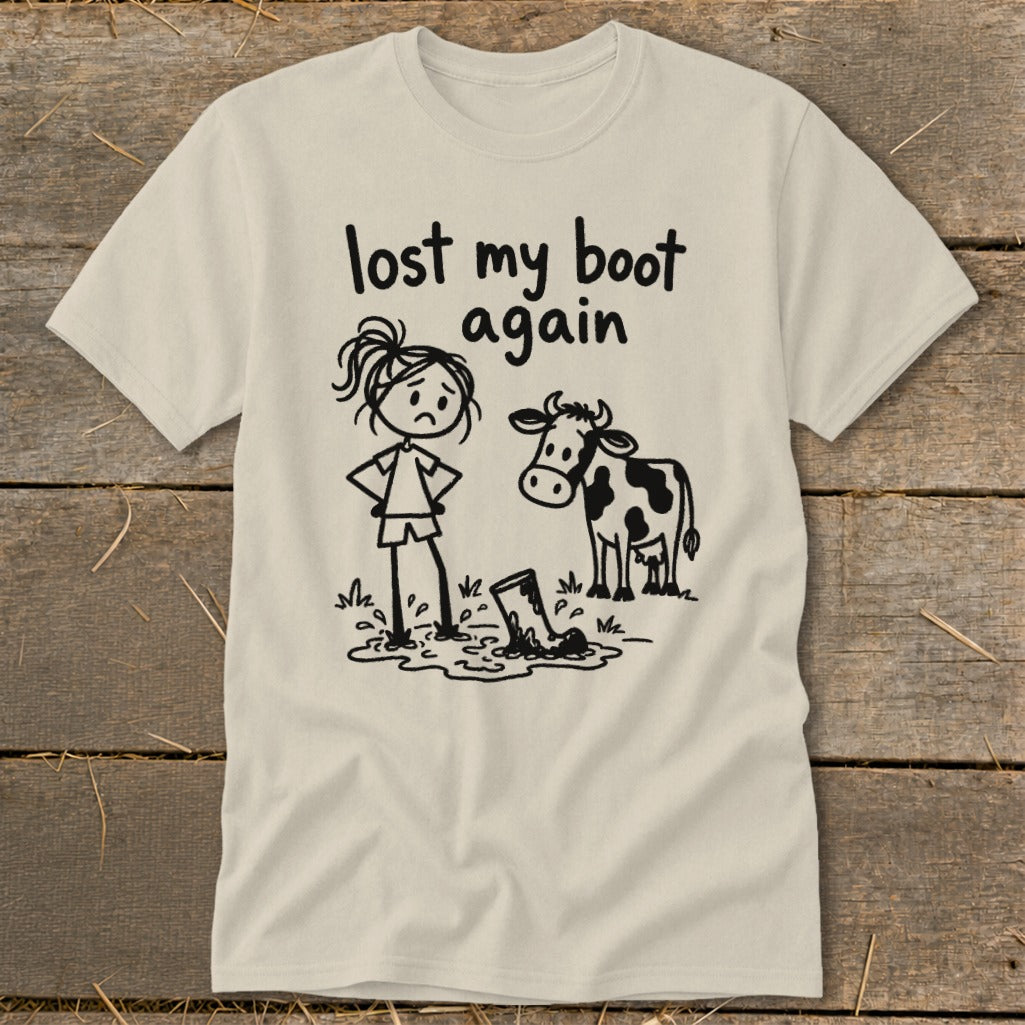 Cream-colored t-shirt with a cartoon graphic of a sad girl and a cow looking at a lost boot in a puddle, featuring the text "lost my boot again."