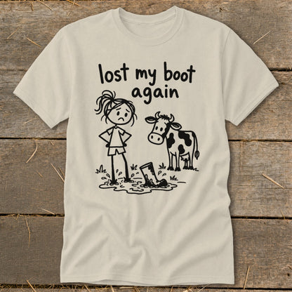 Cream-colored t-shirt with a cartoon graphic of a sad girl and a cow looking at a lost boot in a puddle, featuring the text "lost my boot again."