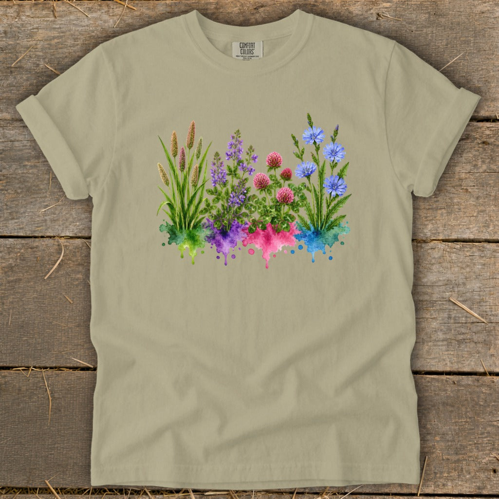 Khaki t-shirt featuring a watercolor graphic of grasses, purple, pink, and blue flowers over colored splatters.