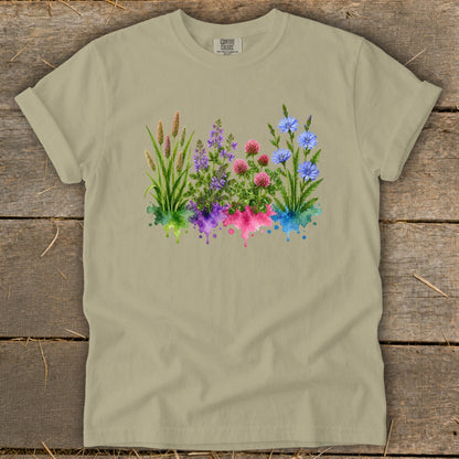 Khaki t-shirt featuring a watercolor graphic of grasses, purple, pink, and blue flowers over colored splatters.