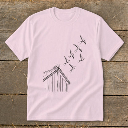 A light pink t-shirt features a black line drawing of birds flying from a rustic wooden cabin roof.