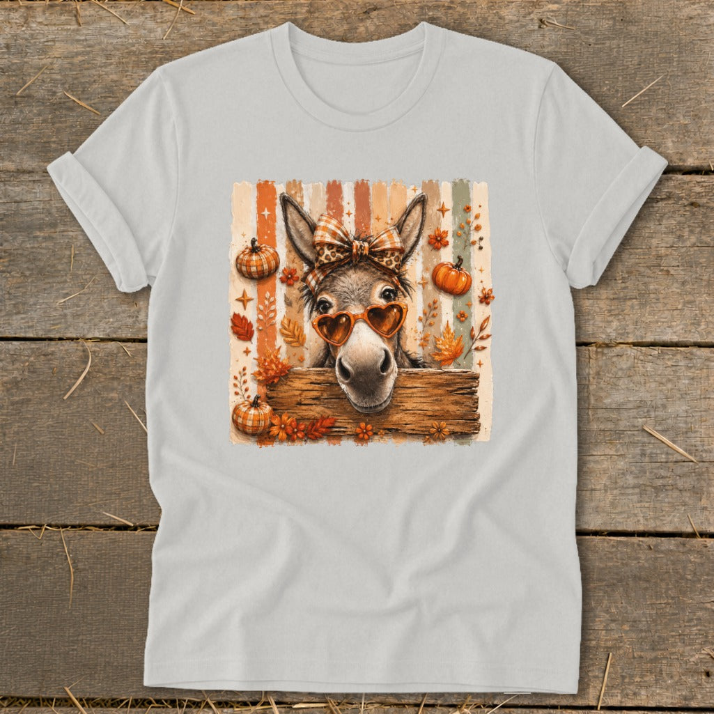 Donkey wearing heart sunglasses and bow peeking over wooden fence with pumpkins and leaves.