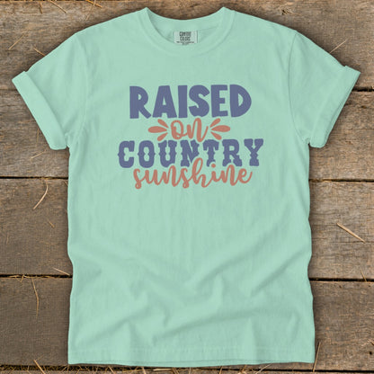 Light green t-shirt with "Raised On Country Sunshine" text in blue and coral.