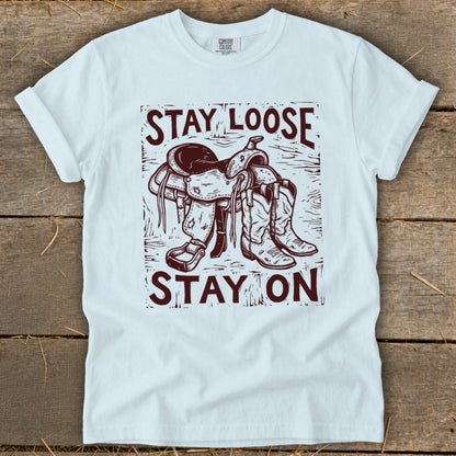 Light blue t-shirt with a graphic of a saddle and cowboy boots and the text "STAY LOOSE STAY ON".