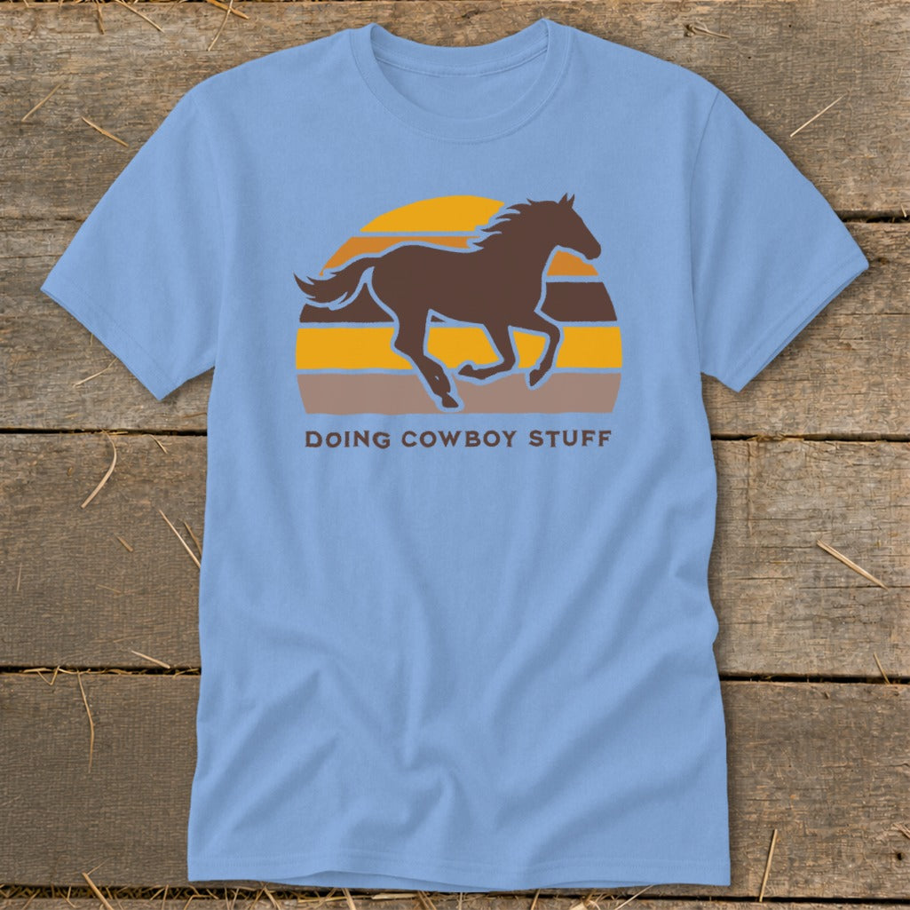 Light blue t-shirt with a brown horse silhouette over a striped sunset and text "DOING COWBOY STUFF".