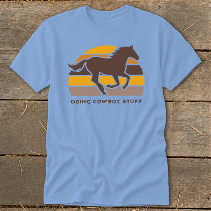Light blue t-shirt with a brown horse silhouette over a striped sunset and text "DOING COWBOY STUFF".