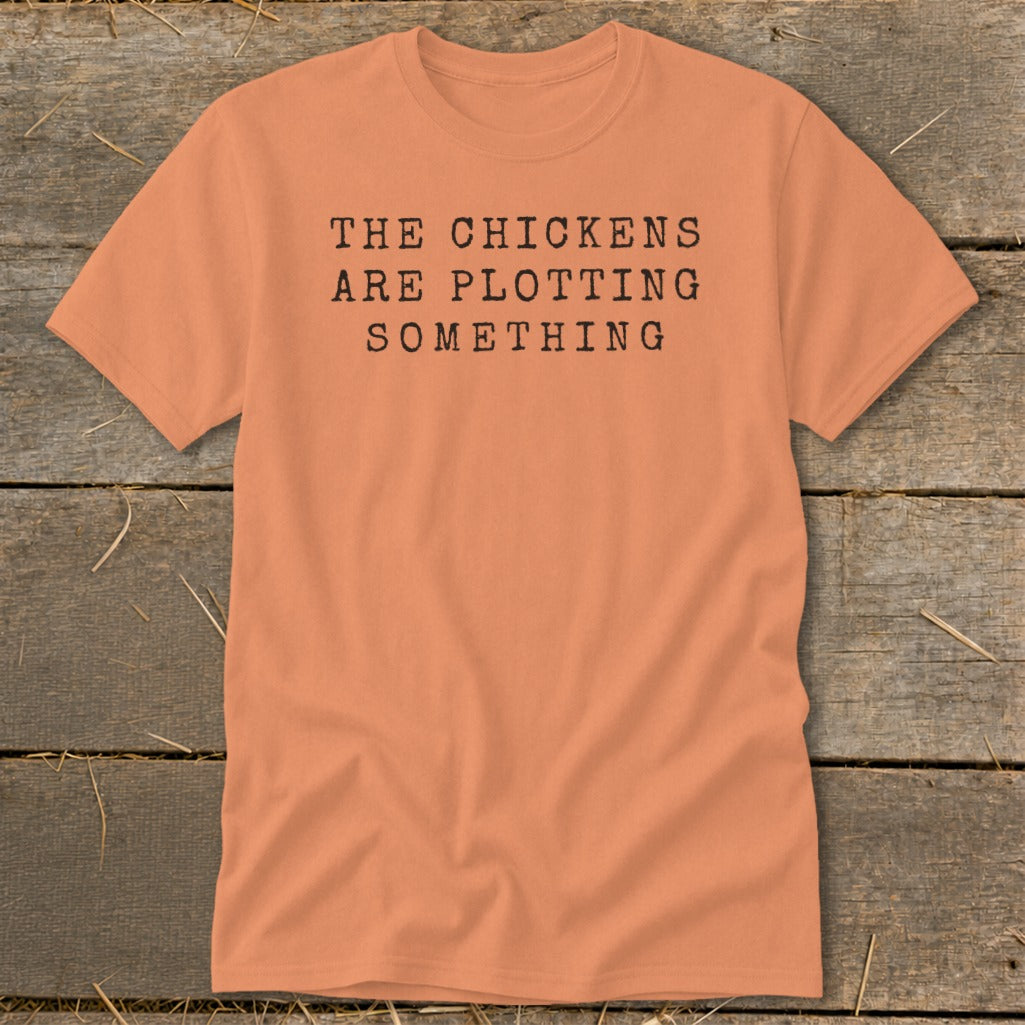 Peach t-shirt with black typewriter text that reads "THE CHICKENS ARE PLOTTING SOMETHING".
