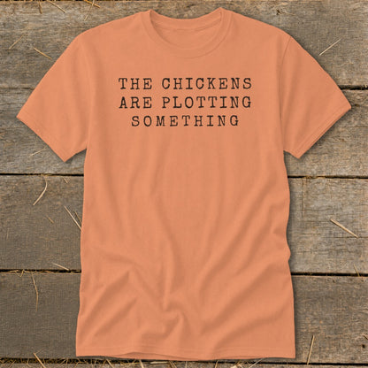 Peach t-shirt with black typewriter text that reads "THE CHICKENS ARE PLOTTING SOMETHING".