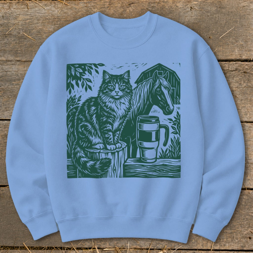 Light blue sweatshirt with green linocut design of cat, horse, and mug on a wooden surface.