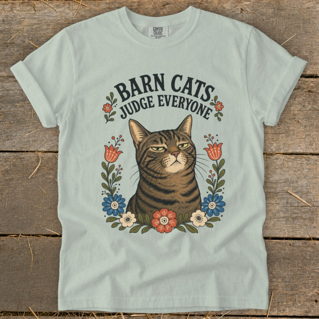 Light blue t-shirt with a tabby cat illustration surrounded by flowers and text "Barn Cats Judge Everyone."