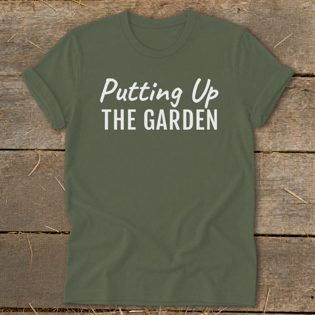 Green t-shirt with white text "Putting Up THE GARDEN" on a wooden background.