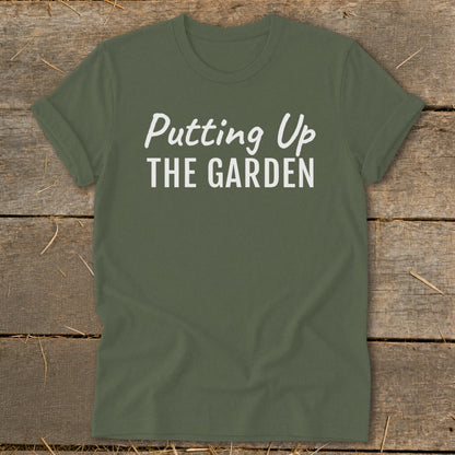 Green t-shirt with white text "Putting Up THE GARDEN" on a wooden background.