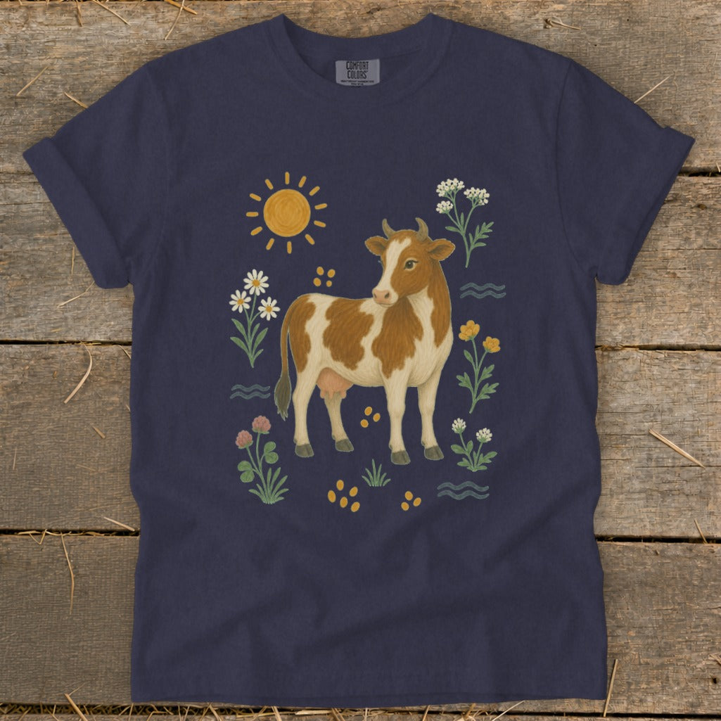 Brown and white cow surrounded by flowers, sun, and waves on a navy t-shirt.