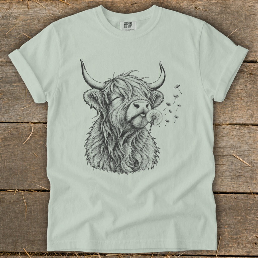 T-shirt featuring a sketch of a Highland cow blowing a dandelion.
