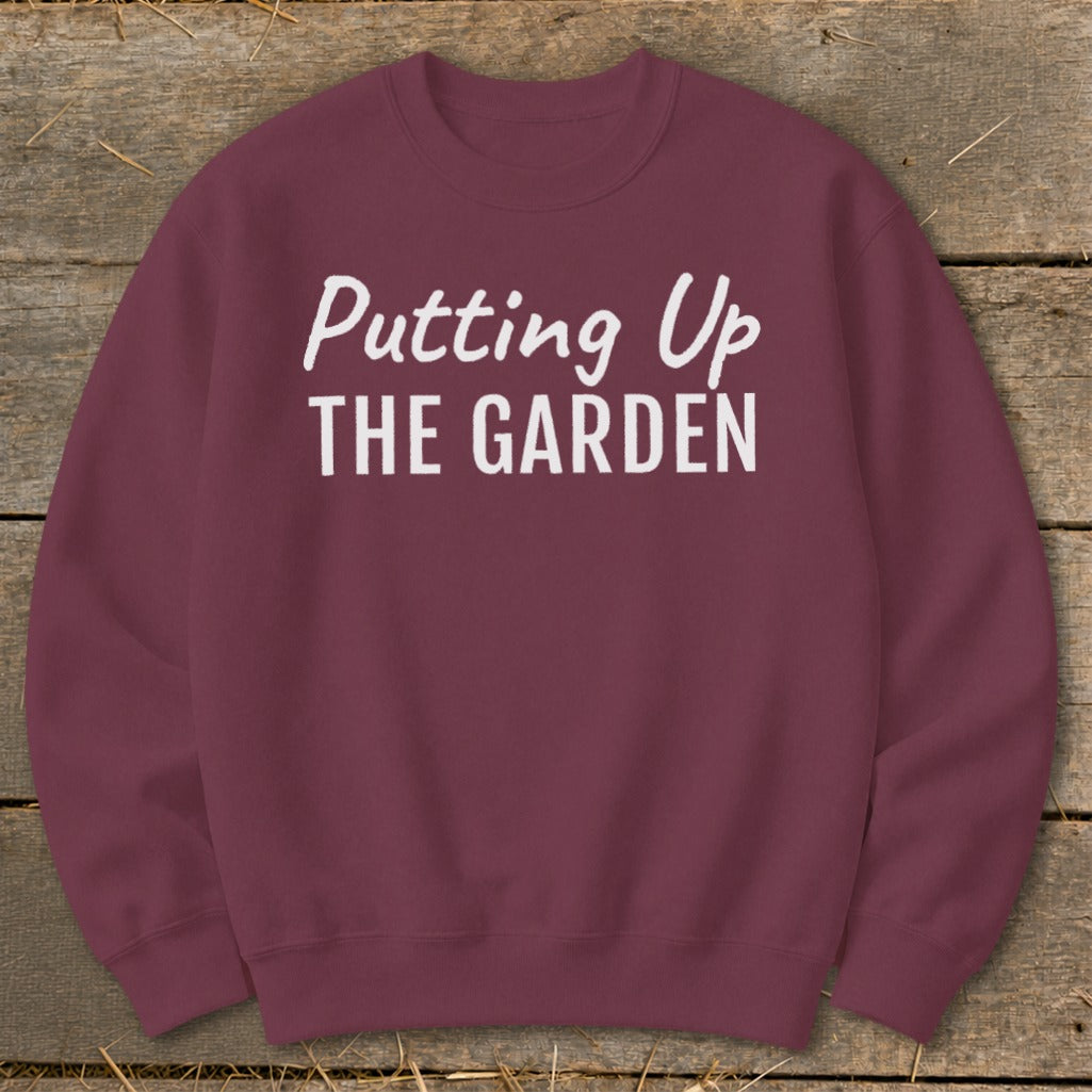 Maroon sweatshirt with white text "Putting Up THE GARDEN" on a wooden background.