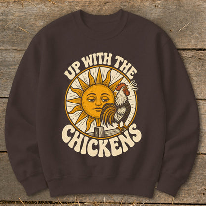 Dark brown crewneck sweatshirt featuring a graphic of a rooster crowing on a fence before a sleepy sun and the text "UP WITH THE CHICKENS".