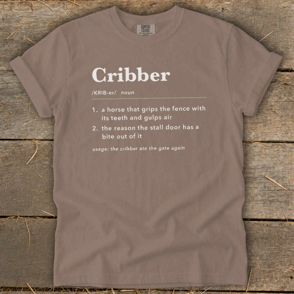 A brown t-shirt featuring white text that defines the word cribber lying on a rustic wooden surface.