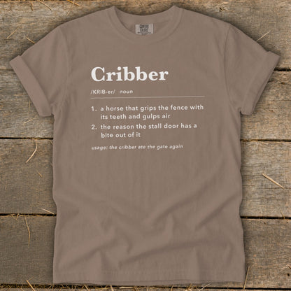 A brown t-shirt featuring white text that defines the word cribber lying on a rustic wooden surface.