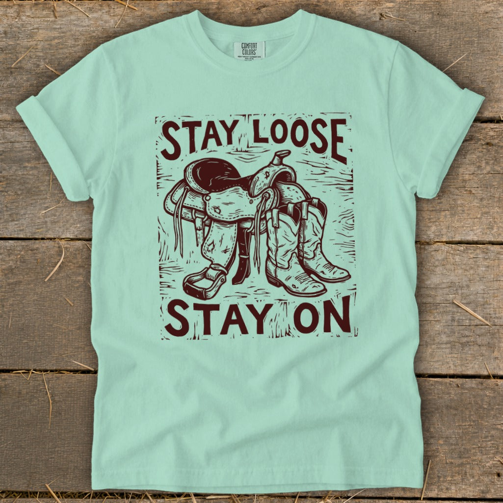 Light blue t-shirt with maroon graphic of a saddle and boots with text "STAY LOOSE STAY ON."