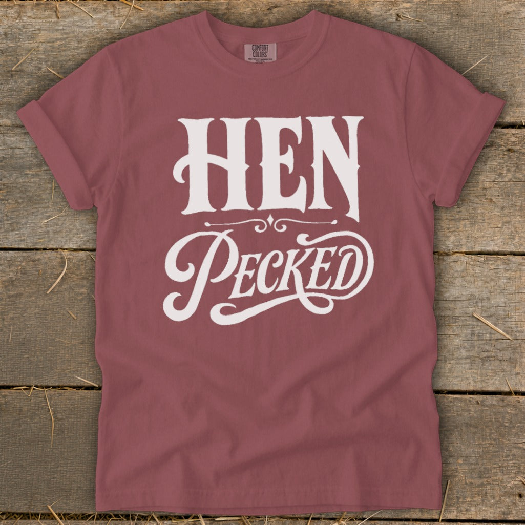 Rose-colored t-shirt with white text "HEN Pecked" on a wooden background.