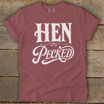 Rose-colored t-shirt with white text "HEN Pecked" on a wooden background.