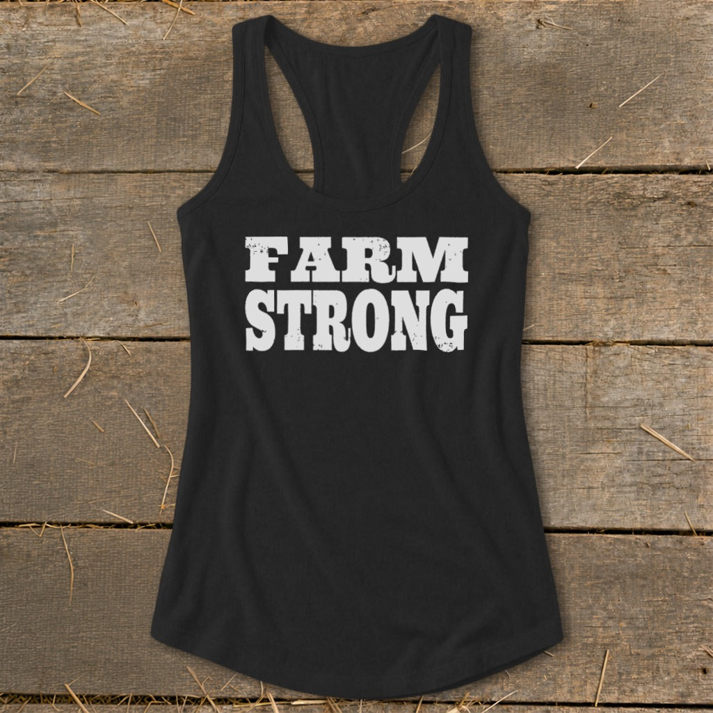 Black women's racerback tank top featuring white distressed text that reads "FARM STRONG" on a rustic wooden background.