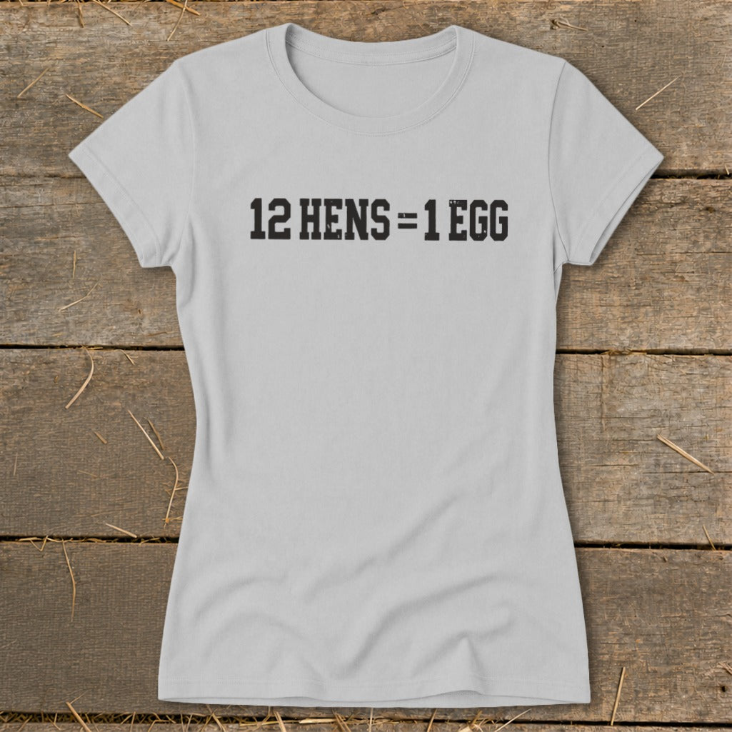 A light gray women's t-shirt printed with the black text 12 HENS = 1 EGG, laid flat on a rustic wooden surface.