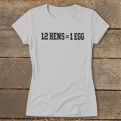 A light gray women's t-shirt printed with the black text 12 HENS = 1 EGG, laid flat on a rustic wooden surface.