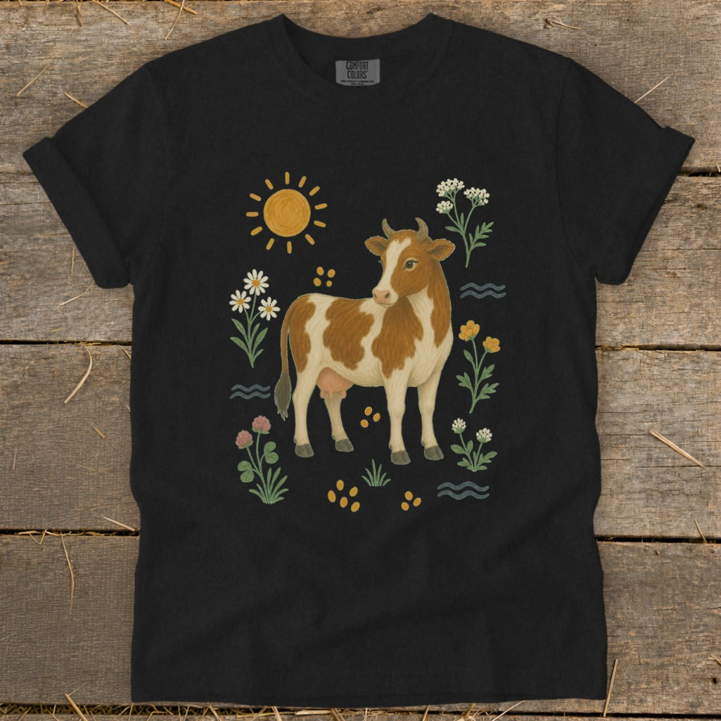Black t-shirt with brown and white cow illustration surrounded by flowers and sun.
