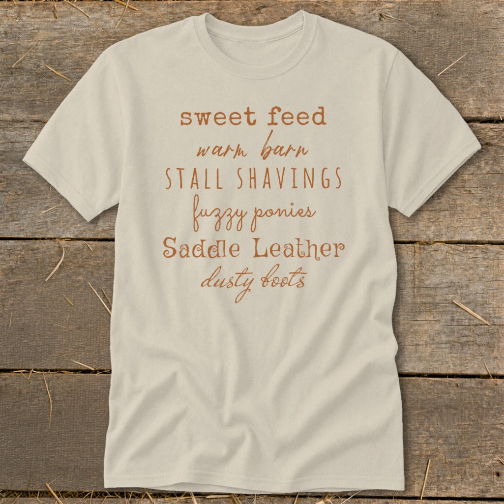 Light-colored t-shirt with text: "sweet feed warm barn STALL SHAVINGS fuzzy ponies Saddle Leather dusty boots."
