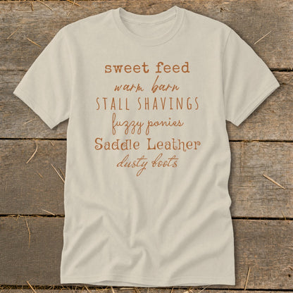 Light-colored t-shirt with text: "sweet feed warm barn STALL SHAVINGS fuzzy ponies Saddle Leather dusty boots."