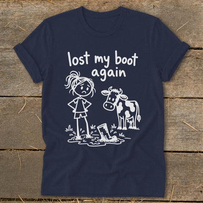 Navy blue t-shirt featuring a white stick figure girl looking at a boot stuck in a mud puddle next to a cow and the text lost my boot again