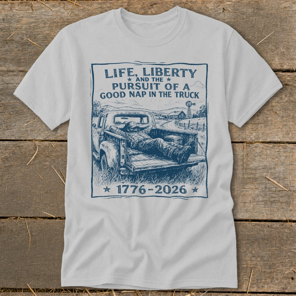T-shirt graphic: "Life, Liberty and the Pursuit of a Good Nap in the Truck" with a man sleeping in a truck bed, 1776-2026.
