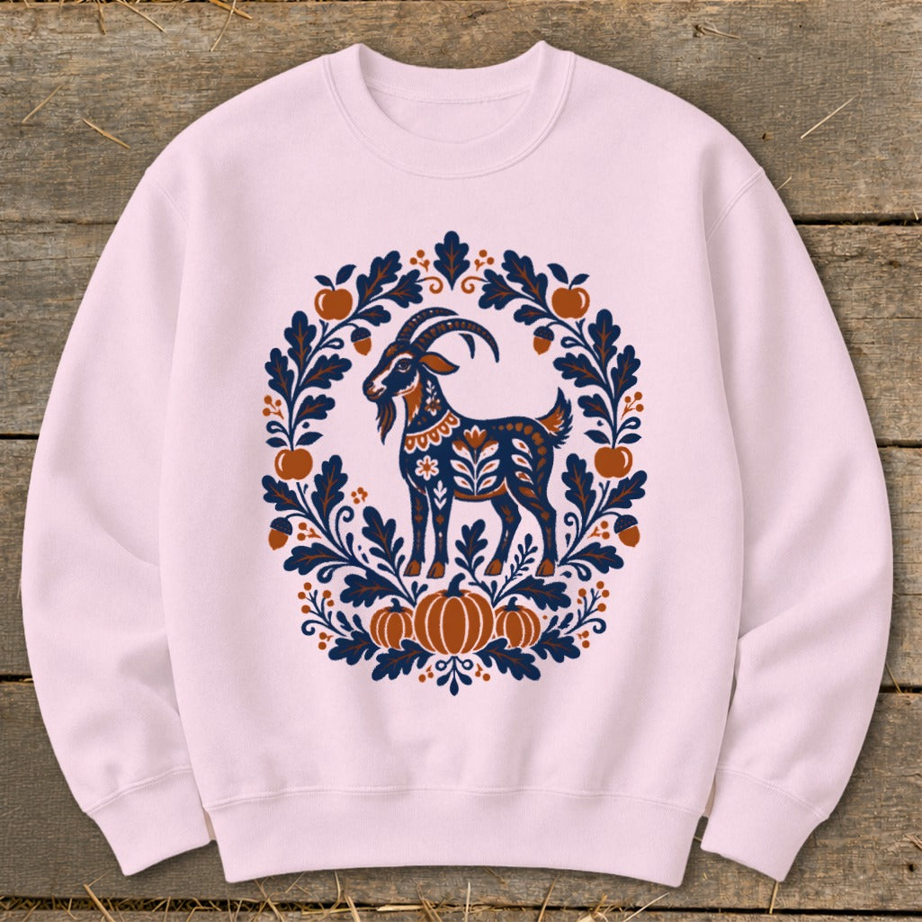 A light pink crewneck sweatshirt features a blue and orange folk-art goat graphic surrounded by pumpkins, apples, and leaves.