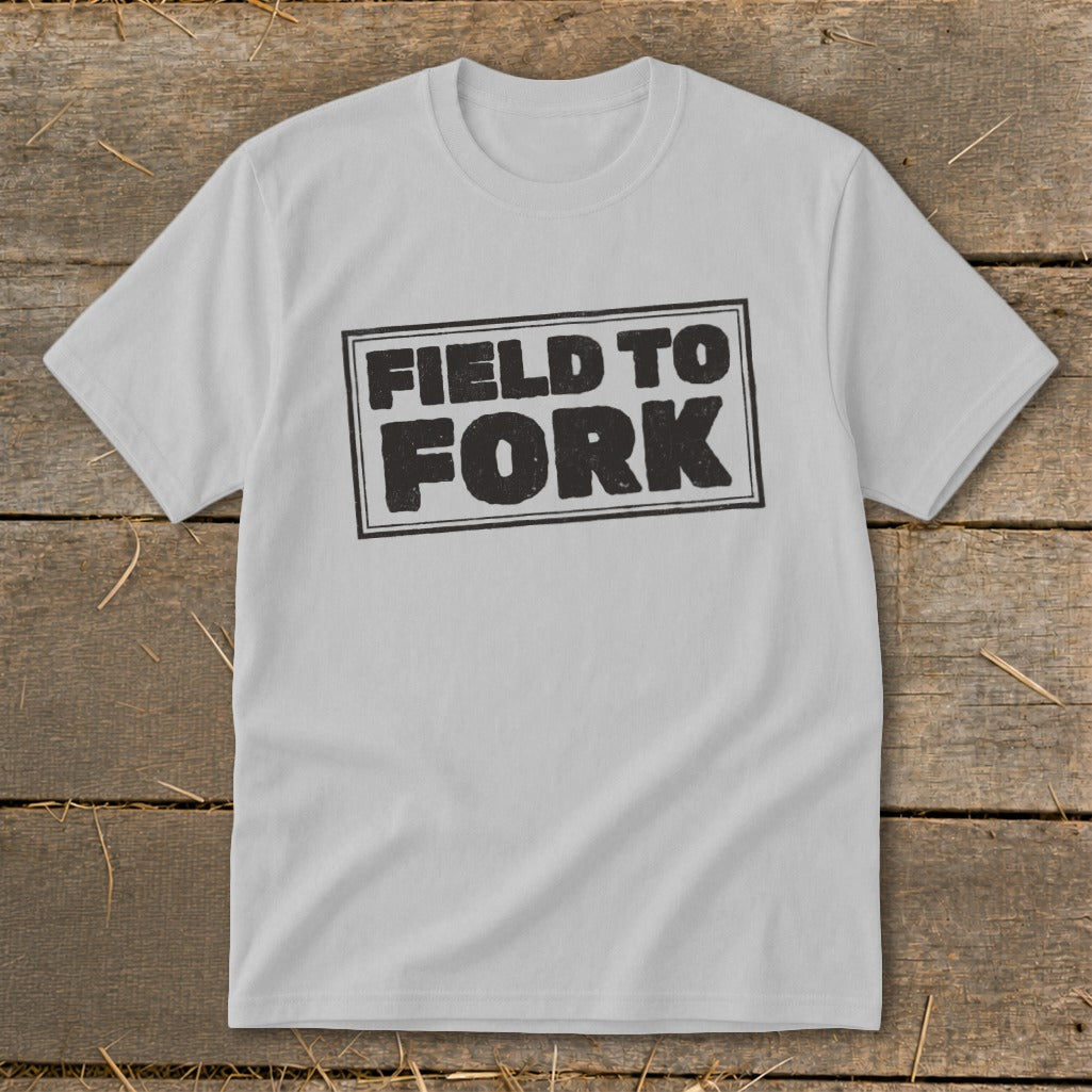 Light gray short-sleeve t-shirt with a rectangular black graphic reading FIELD TO FORK laid on wood.