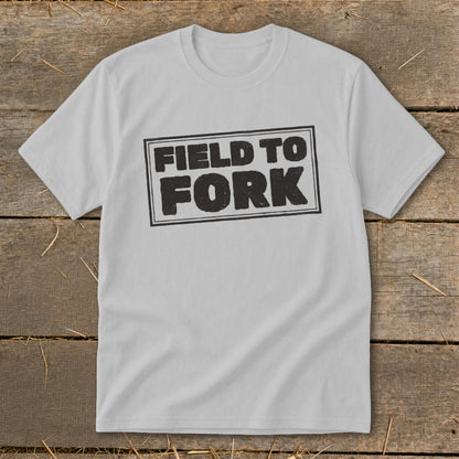 Light gray short-sleeve t-shirt with a rectangular black graphic reading FIELD TO FORK laid on wood.
