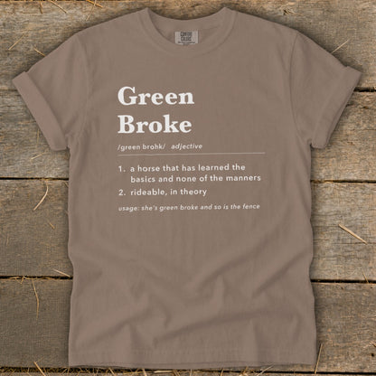 A brown t-shirt featuring white text that defines the adjective Green Broke on a rustic wooden background.