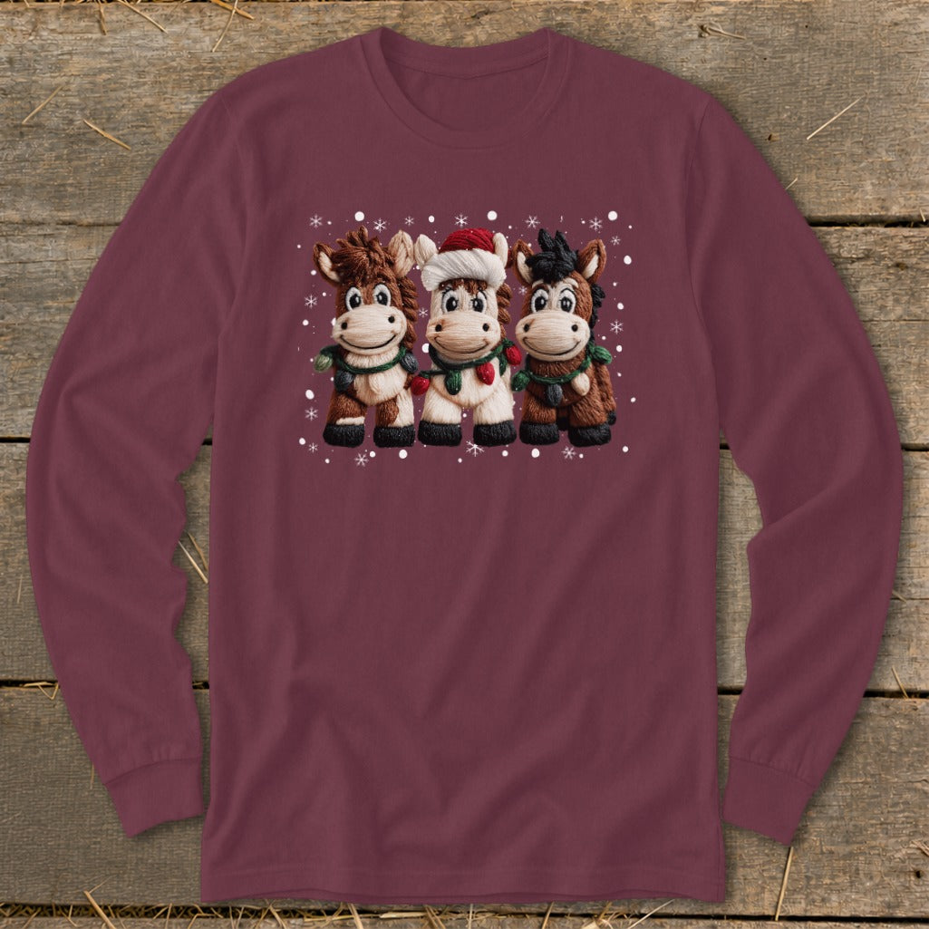 Three cartoon horses wearing Christmas lights and scarves on a maroon long-sleeve shirt.