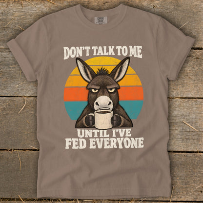 Gray t-shirt featuring a grumpy donkey drinking from a mug with text that reads don't talk to me until i've fed everyone.