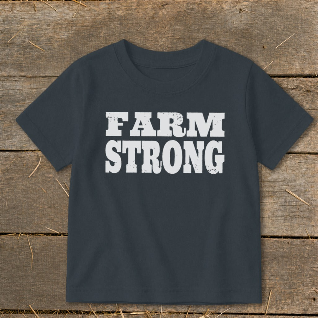 Dark gray short-sleeved children's t-shirt featuring the distressed white text FARM STRONG laid flat on a wooden background.