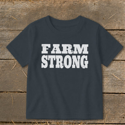 Dark gray short-sleeved children's t-shirt featuring the distressed white text FARM STRONG laid flat on a wooden background.