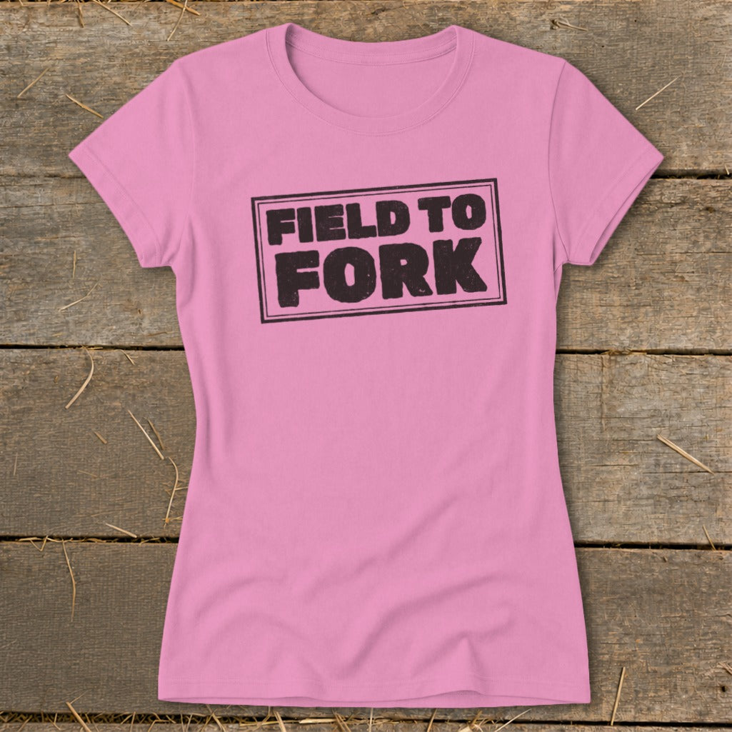 A pink women's t-shirt rests on a rustic wooden background, featuring the black text Field to Fork.