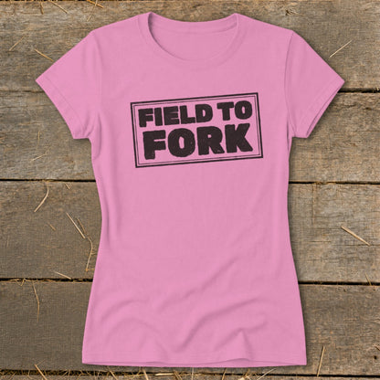 A pink women's t-shirt rests on a rustic wooden background, featuring the black text Field to Fork.